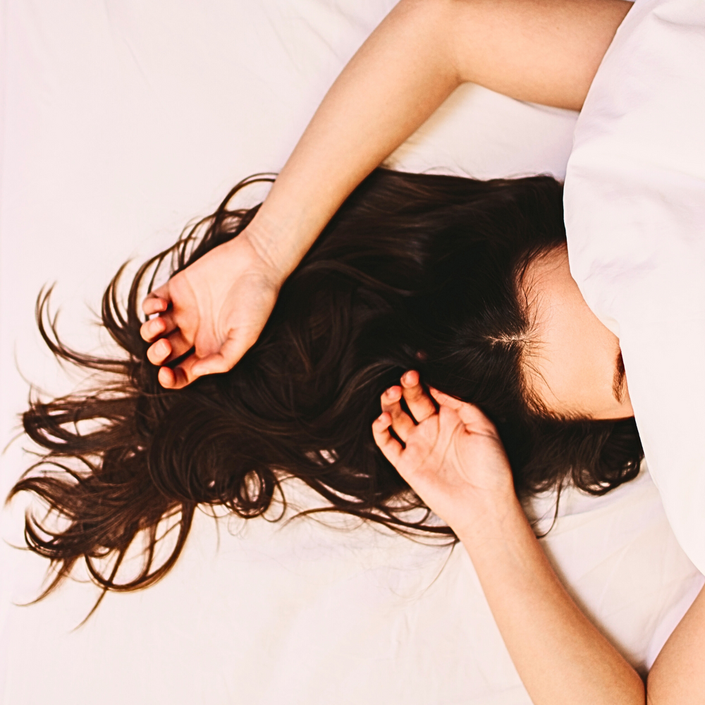Top-down view of a woman lying in bed with long dark hair spread out, partially covered by a white blanket—symbolizing rest, relaxation, and deep sleep